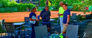 Vier Personen stehen auf einer Terrasse mit Metalltischen und -st&uuml;hlen, im Hintergrund ist ein Tennisplatz zu sehen.