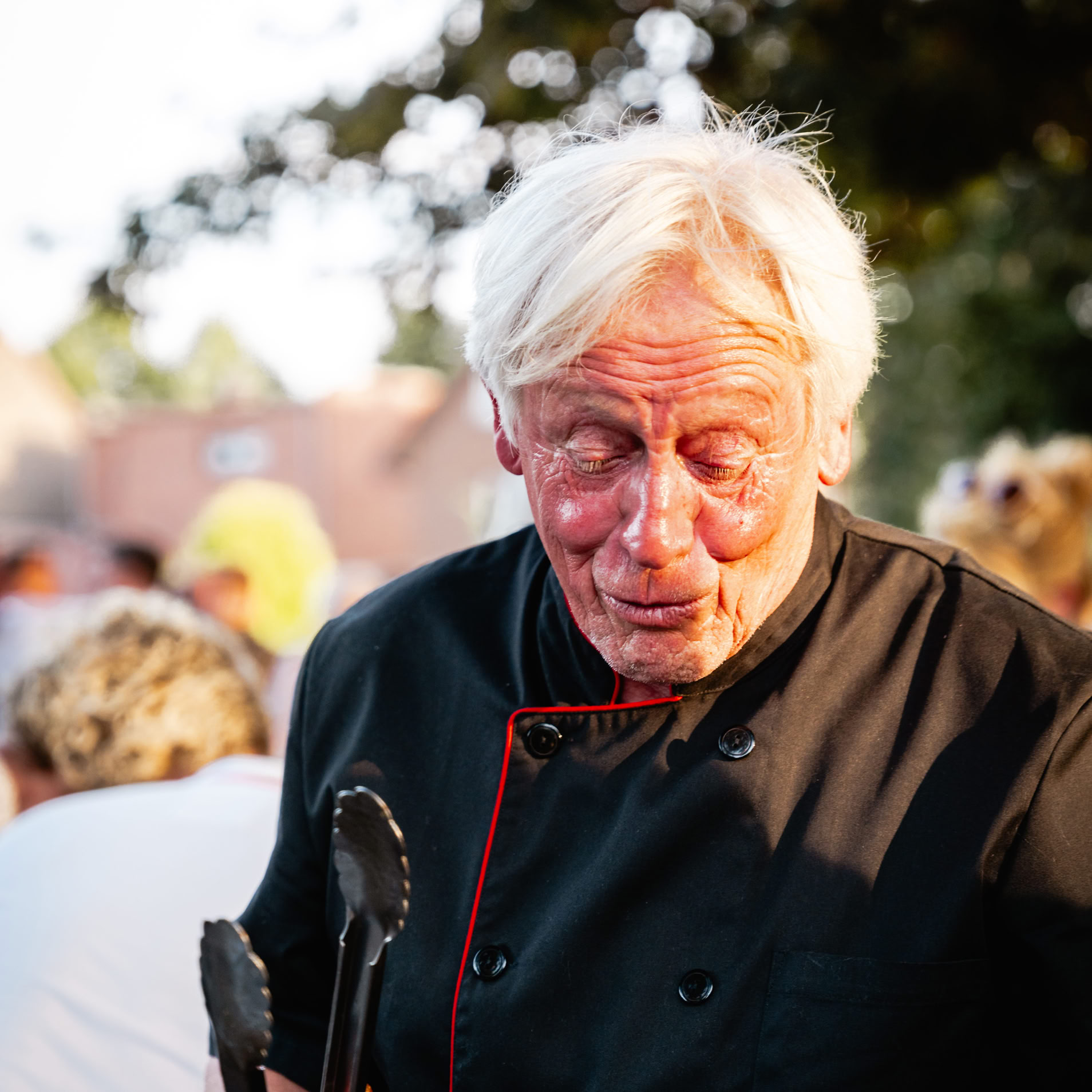 &Auml;lterer Mann mit wei&szlig;em Haar in schwarzer Kochjacke h&auml;lt Grillzange, im Freien mit unscharfem Hintergrund.