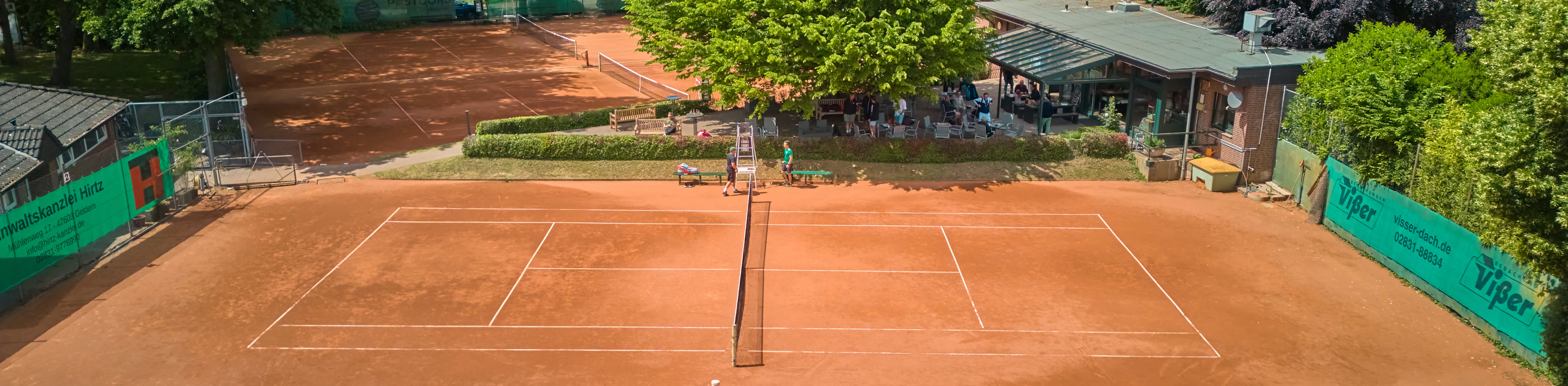 Tennisplatz mit rotem Sandbelag, umgeben von B&auml;umen und einem Geb&auml;ude im Hintergrund.