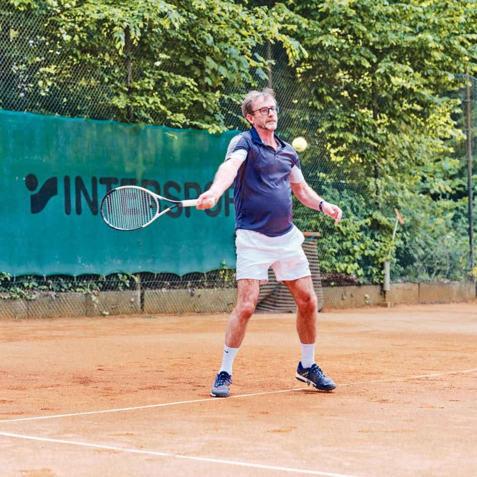 Eine Person spielt Tennis auf einem Sandplatz, umgeben von B&auml;umen, mit einem gr&uuml;nen Zaun im Hintergrund.