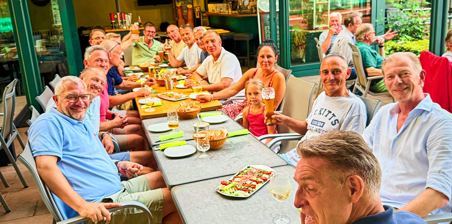 Eine Gruppe von Menschen sitzt an einem langen Tisch im Freien, mit Tellern und Gl&auml;sern vor sich. Im Hintergrund ist ein Geb&auml;ude mit offenen T&uuml;ren und Fenstern zu sehen.