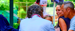 Eine Gruppe von Menschen sitzt an einem Tisch im Freien, umgeben von B&auml;umen und Pflanzen.