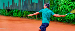 Person spielt Tennis auf einem Sandplatz, umgeben von gr&uuml;ner Vegetation und einem Zaun im Hintergrund.