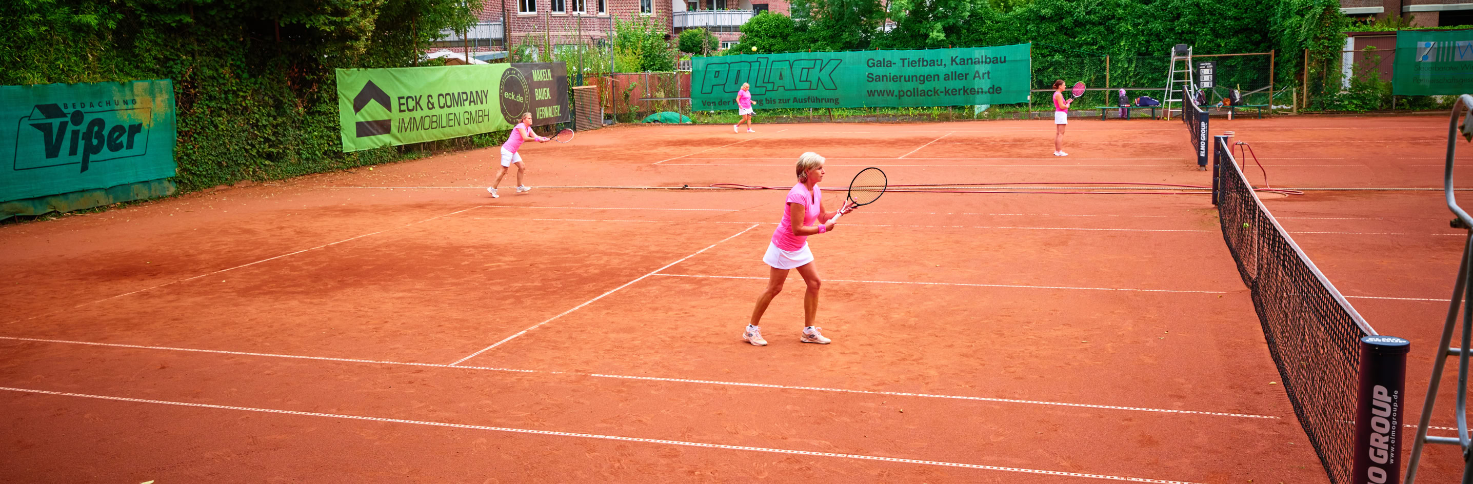 Tennisplatz mit mehreren Personen, die Tennis spielen; im Vordergrund liegen Taschen, im Hintergrund sind B&auml;ume und Geb&auml;ude sichtbar.