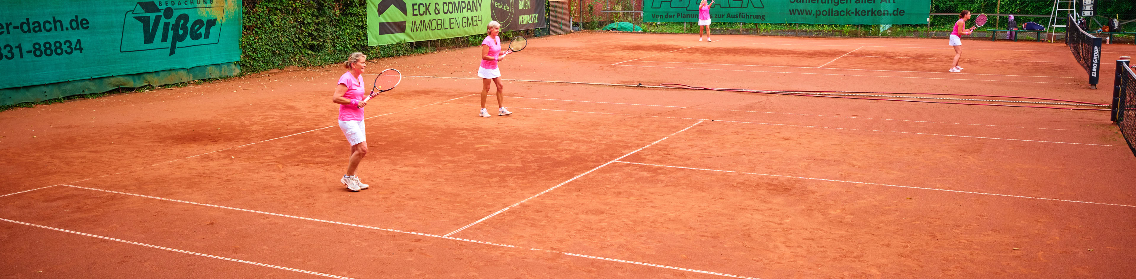 Tennisplatz mit vier Personen, die Tennis spielen; umgeben von B&auml;umen und Geb&auml;uden im Hintergrund.