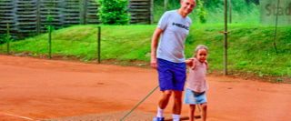 Ein Erwachsener und ein Kind stehen auf einem Tennisplatz mit rotem Sand, umgeben von einem Zaun und gr&uuml;ner Vegetation.
