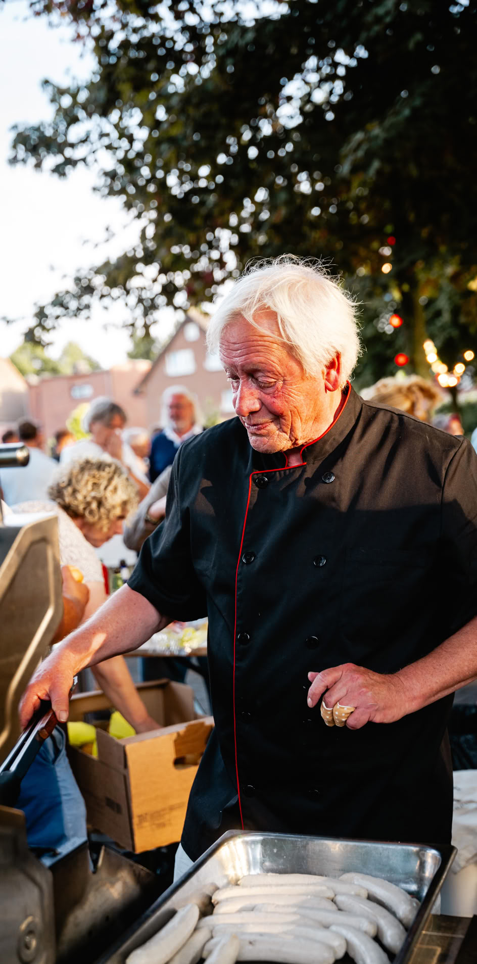 &Auml;lterer Mann in schwarzer Kleidung steht drau&szlig;en an einem Grill, umgeben von Menschen.