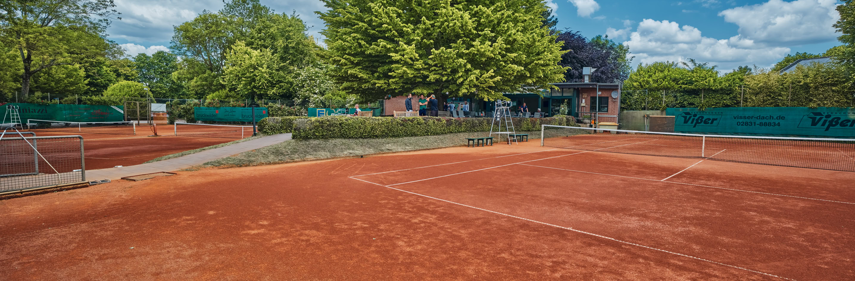 Ein Tennisplatz mit rotem Sandboden, umgeben von B&auml;umen und einem Zaun, unter einem blauen Himmel mit einigen Wolken.