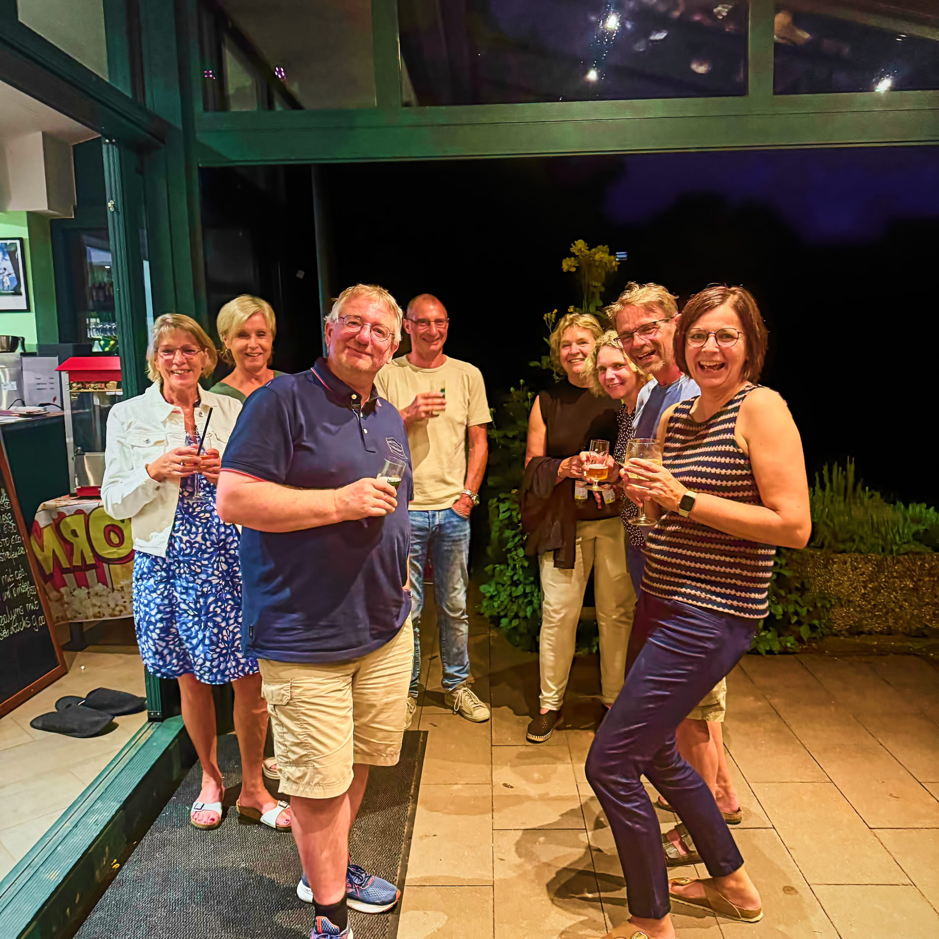 Acht Personen stehen zusammen in einem beleuchteten Raum mit Glasw&auml;nden, einige halten Getr&auml;nke in der Hand. Im Hintergrund ist es dunkel.