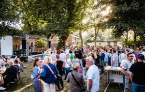 Eine gro&szlig;e Menschenmenge versammelt sich im Freien unter B&auml;umen, einige stehen an einem Tisch, w&auml;hrend andere in Richtung einer B&uuml;hne blicken.