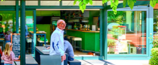 Eine Gruppe von Menschen steht vor einem Geb&auml;ude mit gro&szlig;en Fenstern und gr&uuml;nen Rahmen, umgeben von B&auml;umen und Pflanzen.