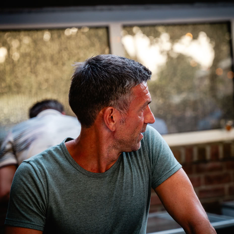 Ein Mann in einem grauen T-Shirt schaut zur Seite, im Hintergrund ist ein Fenster mit einem verschwommenen Ausblick.