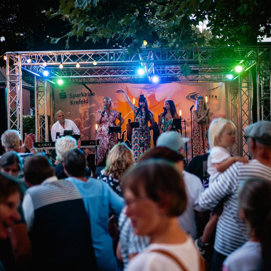 Eine Menschenmenge steht vor einer B&uuml;hne im Freien, auf der mehrere Personen unter bunten Lichtern auftreten.