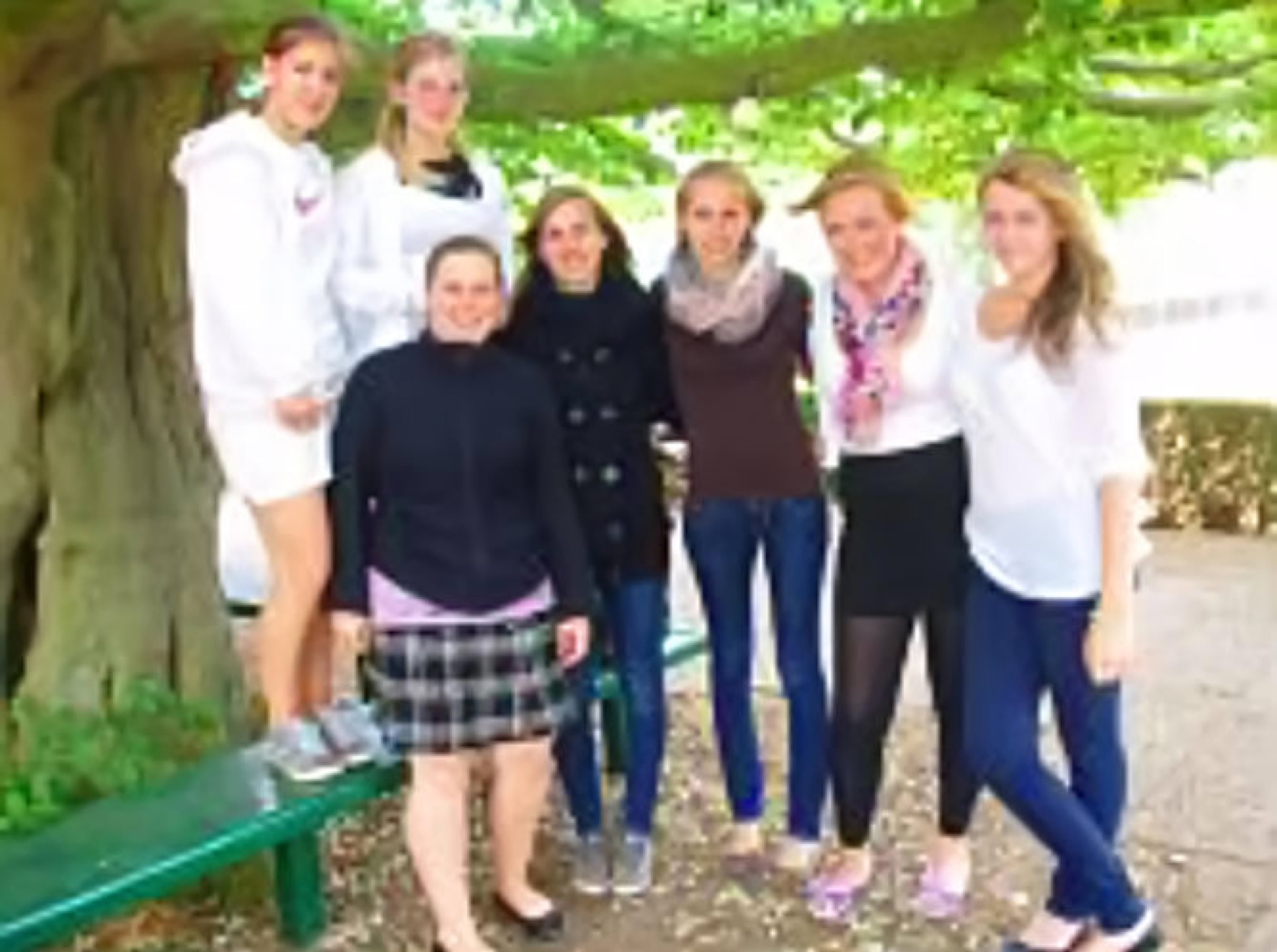 Sieben Personen stehen und sitzen unter einem gro&szlig;en Baum auf einer Bank.