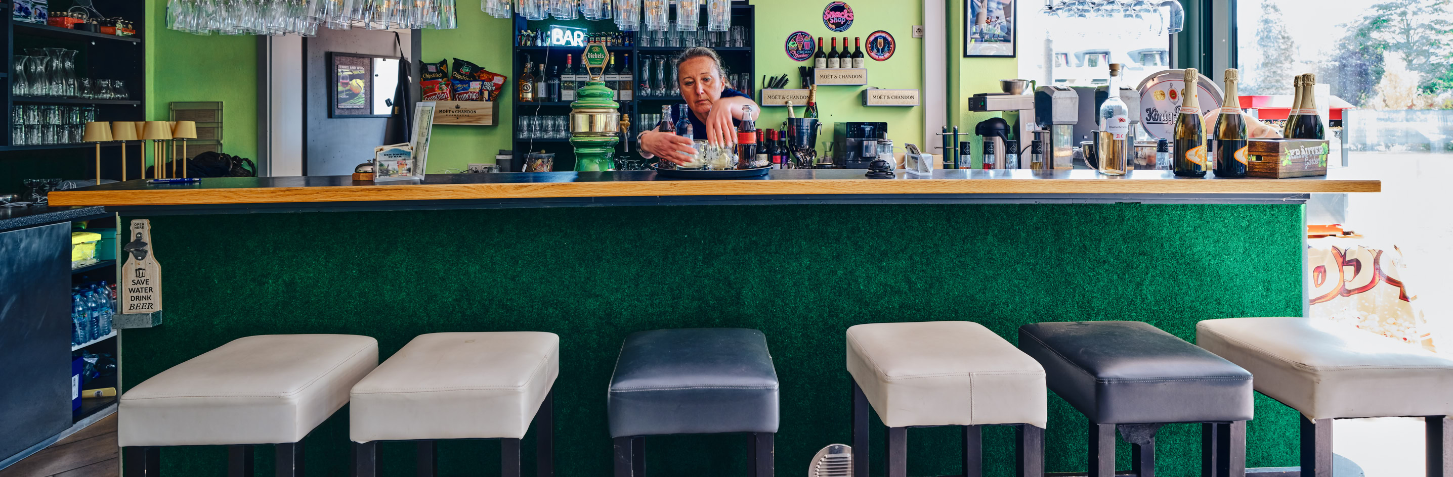 Bar mit gr&uuml;ner Theke, mehreren Hockern und einer Person hinter der Theke. Im Hintergrund sind Regale mit Gl&auml;sern und Flaschen.