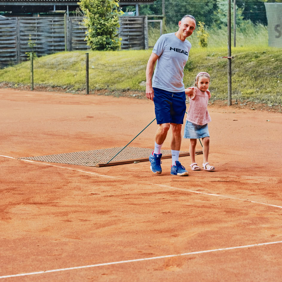 Ein Mann und ein Kind stehen auf einem Tennisplatz aus Sand und halten gemeinsam ein Netz.