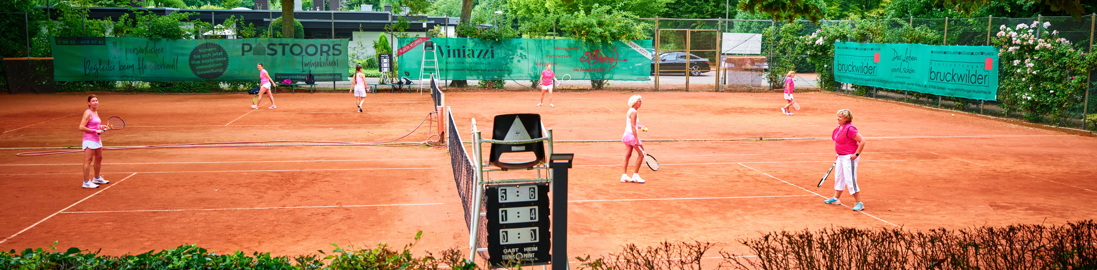 Tennisplatz mit mehreren Personen, die Tennis spielen; im Vordergrund eine Terrasse mit St&uuml;hlen und Tisch, umgeben von B&auml;umen und gr&uuml;nen Hecken.