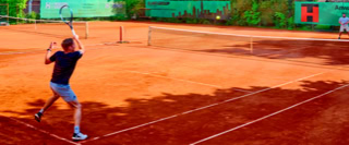 Zwei Personen spielen Tennis auf einem Sandplatz, umgeben von gr&uuml;nen W&auml;nden und B&auml;umen.