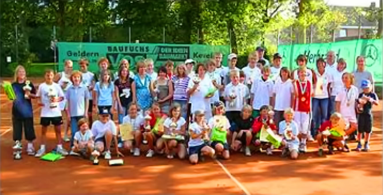 Eine gro&szlig;e Gruppe von Menschen, darunter Kinder und Erwachsene, steht auf einem Tennisplatz. Einige halten Troph&auml;en und Blumenstr&auml;u&szlig;e. Im Hintergrund sind gr&uuml;ne B&auml;ume und ein Tennisnetz sichtbar.