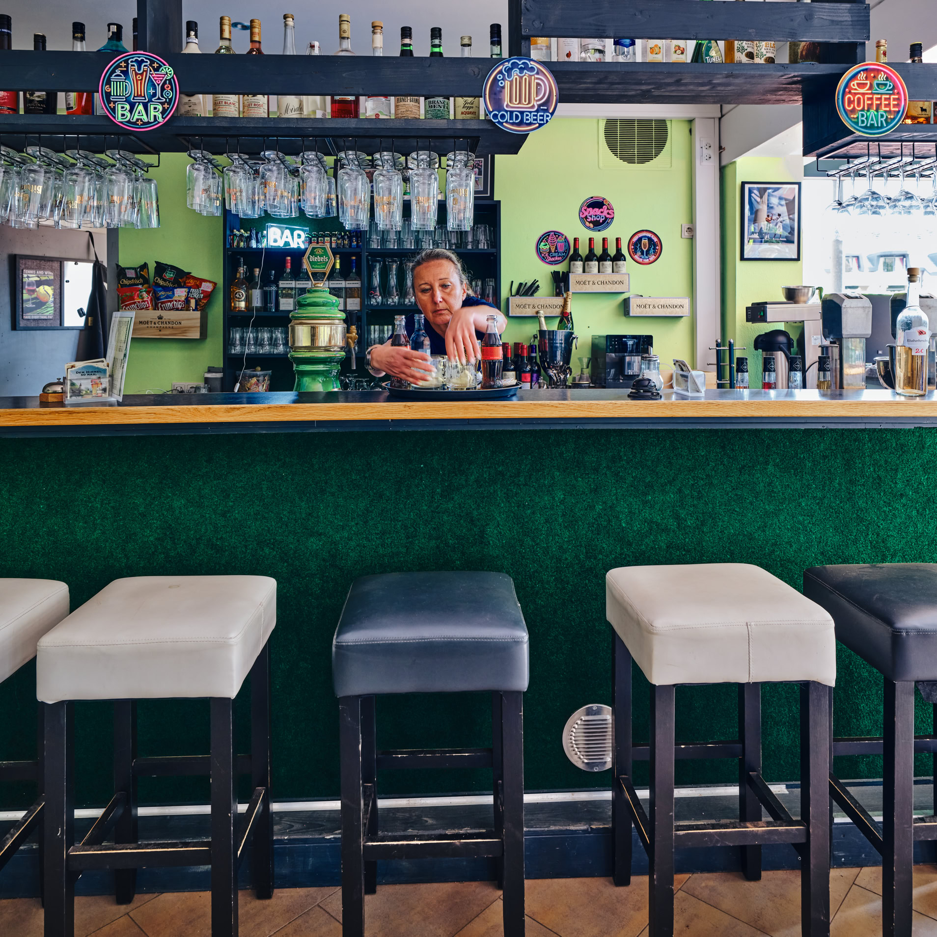 Bar mit gr&uuml;ner Theke, mehreren Hockern und einer Person hinter der Theke. Im Hintergrund sind Regale mit Gl&auml;sern und Flaschen.