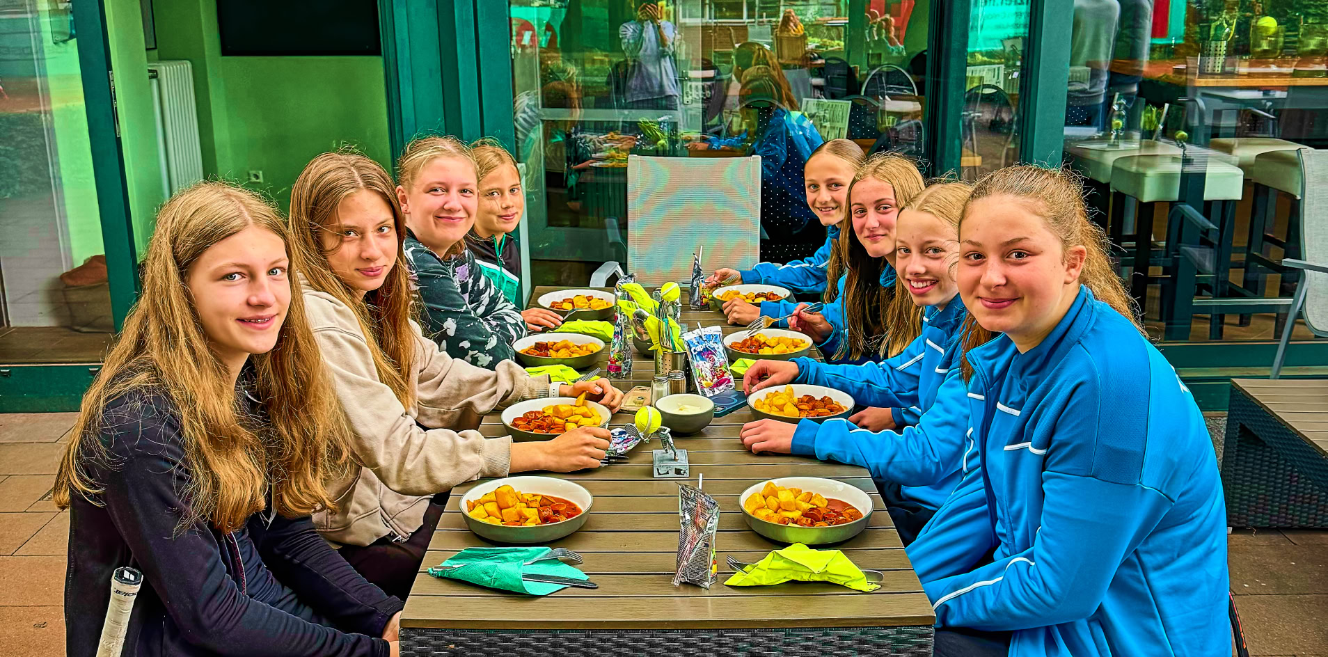 Eine Gruppe von neun Personen sitzt an einem Tisch im Freien und isst. Der Tisch ist mit Tellern und Getr&auml;nken gedeckt. Im Hintergrund ist ein Geb&auml;ude mit Glasfront zu sehen.