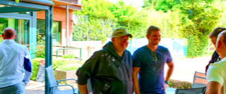 Eine Gruppe von Menschen steht drau&szlig;en vor einem Geb&auml;ude, umgeben von B&auml;umen und Gr&uuml;nfl&auml;chen.