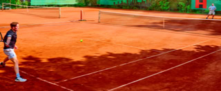 Zwei Personen spielen Tennis auf einem Sandplatz, umgeben von gr&uuml;nen W&auml;nden und B&auml;umen.