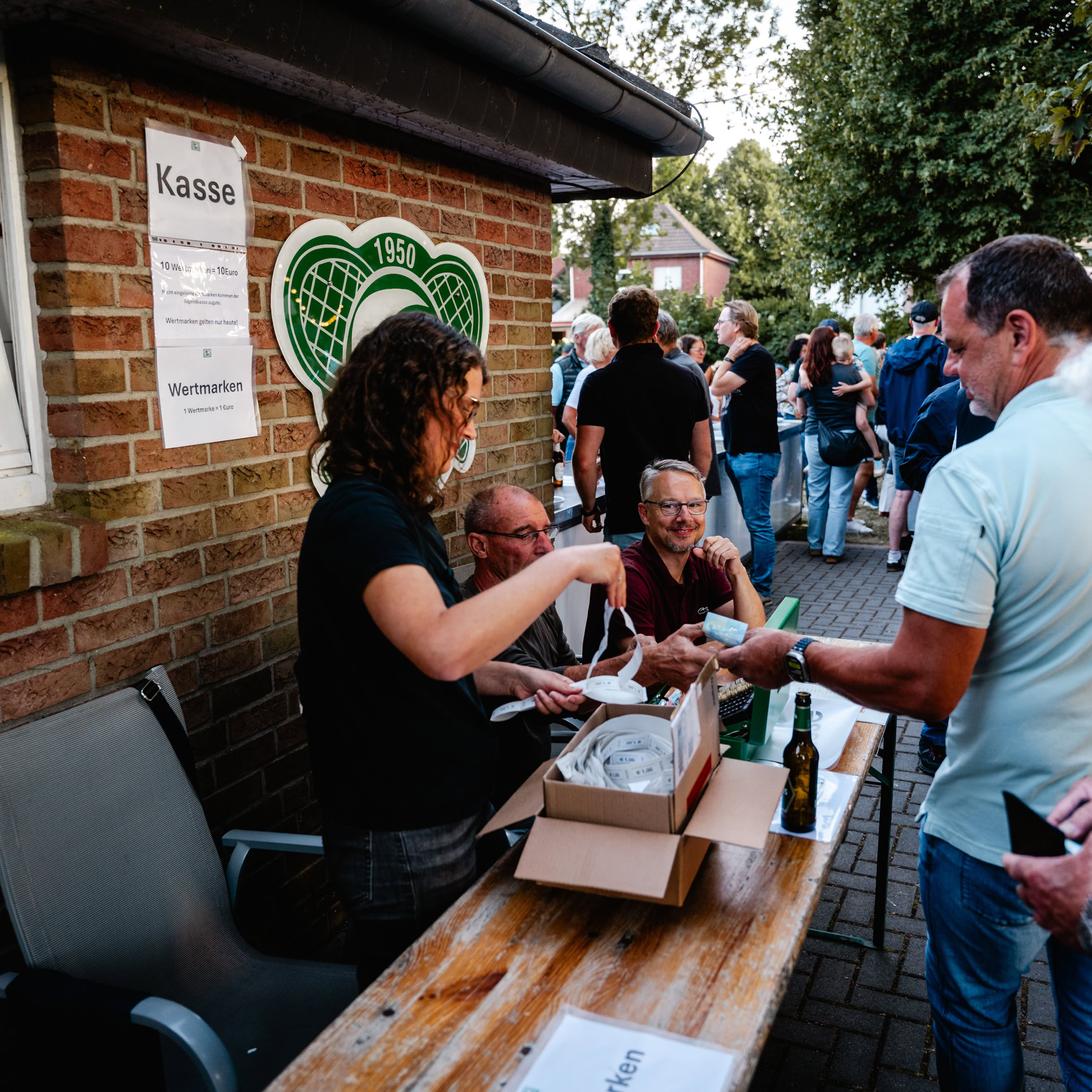 Eine Gruppe von Menschen steht an einem Tisch vor einem Backsteingeb&auml;ude. Eine Person hinter dem Tisch gibt etwas an eine Person davor. Auf dem Tisch stehen eine Schachtel und ein Getr&auml;nk. Im Hintergrund sind weitere Personen und B&auml;ume zu sehen.