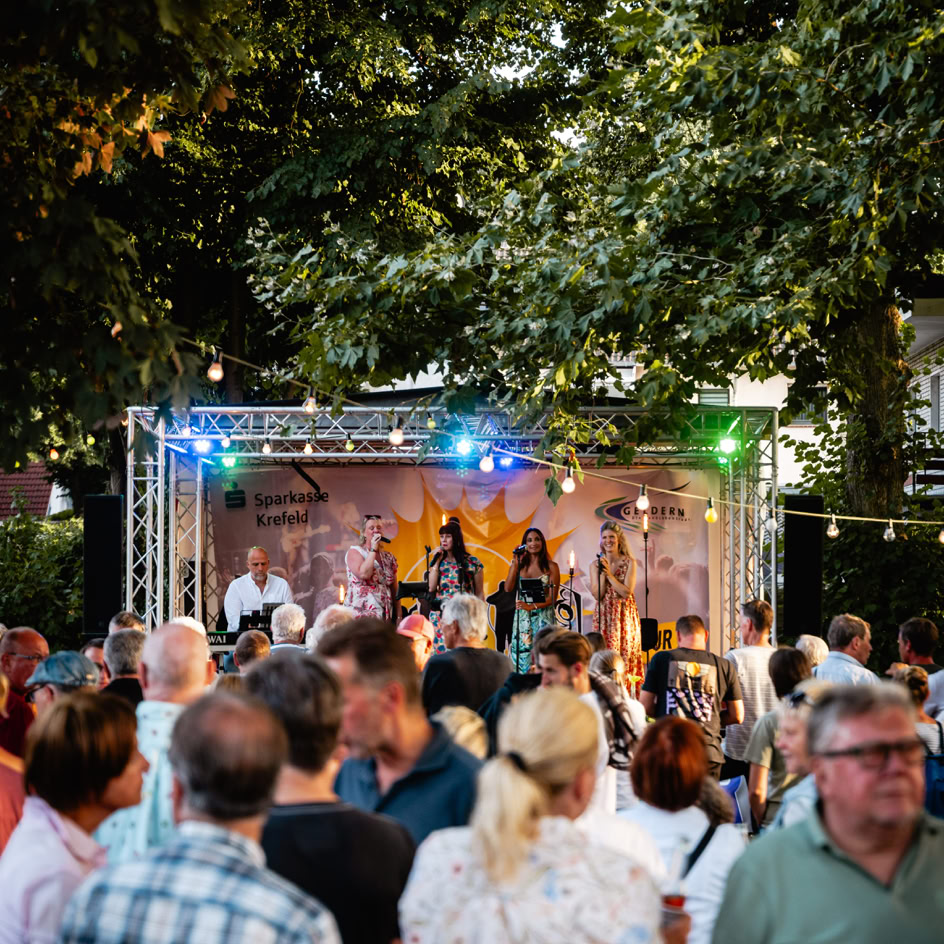 Menschenmenge vor einer B&uuml;hne im Freien mit bunten Lichtern, umgeben von B&auml;umen.