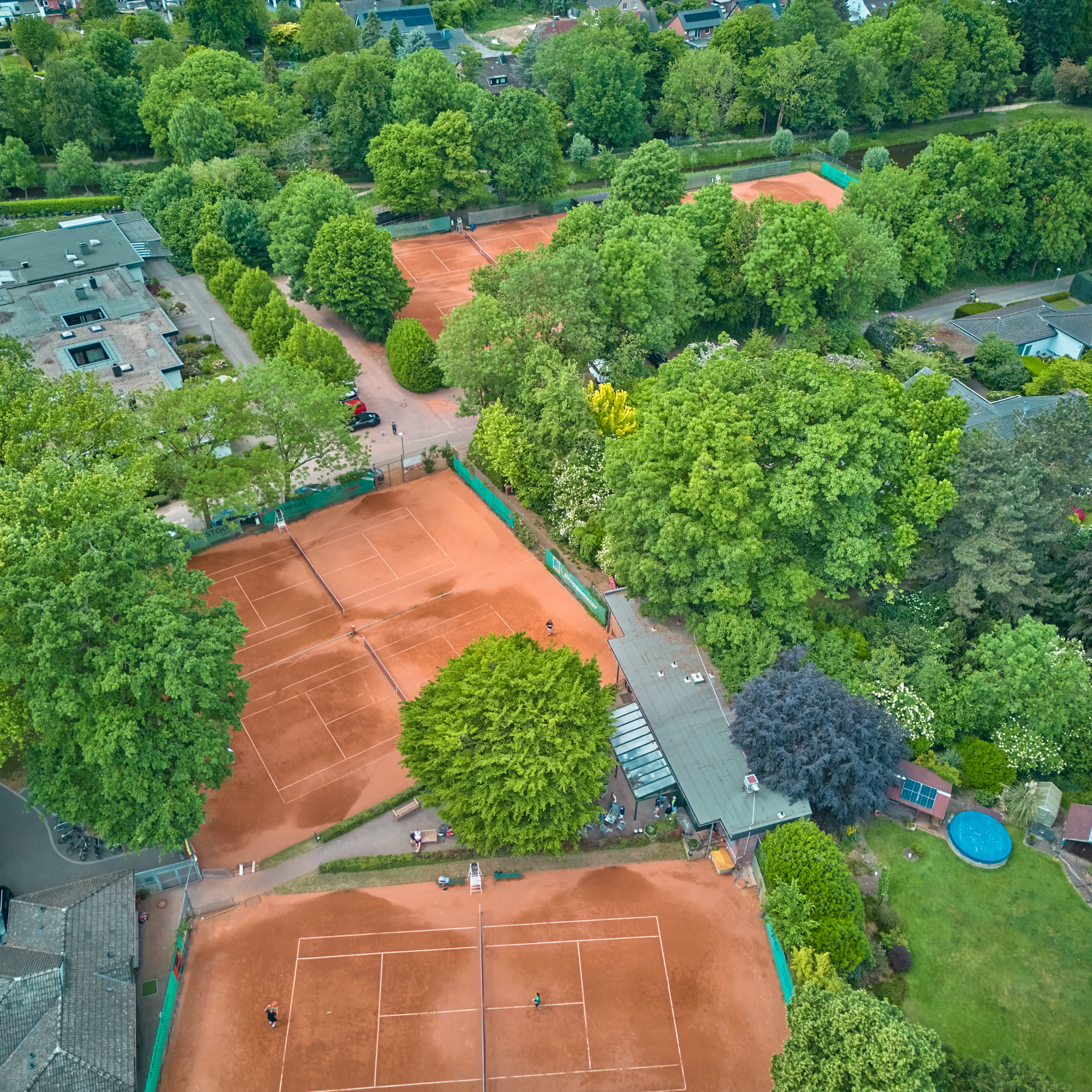 Luftaufnahme von mehreren Tennispl&auml;tzen mit rotem Sand, umgeben von B&auml;umen und Geb&auml;uden.