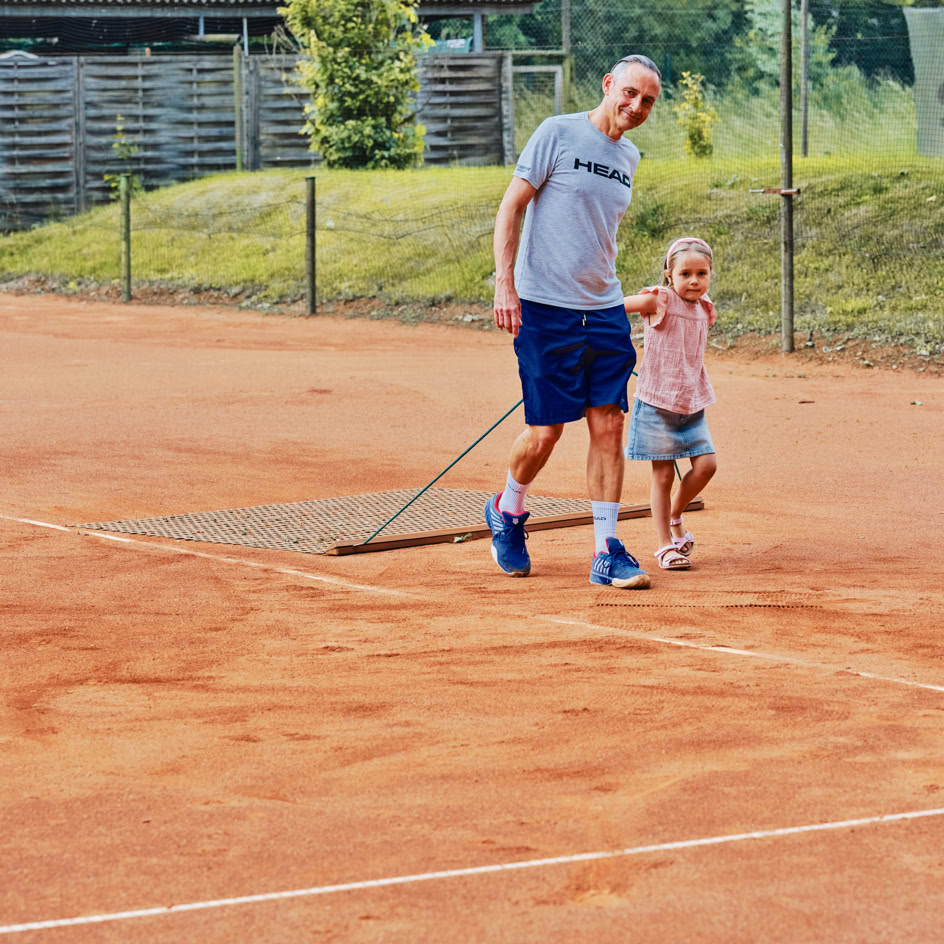Ein Mann und ein Kind ziehen ein Netz &uuml;ber einen Sandtennisplatz.