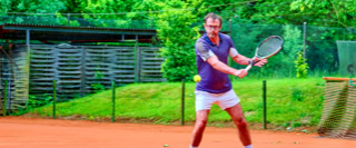 Eine Person spielt Tennis auf einem Sandplatz, umgeben von gr&uuml;ner Vegetation und einem Zaun im Hintergrund.