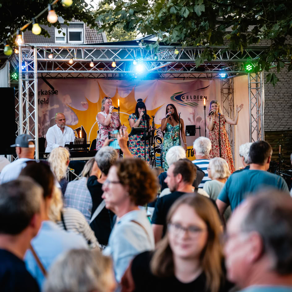 Eine Gruppe von Menschen steht vor einer B&uuml;hne im Freien, auf der Musiker unter bunten Lichtern auftreten. Im Hintergrund sind B&auml;ume und Lichterketten zu sehen.