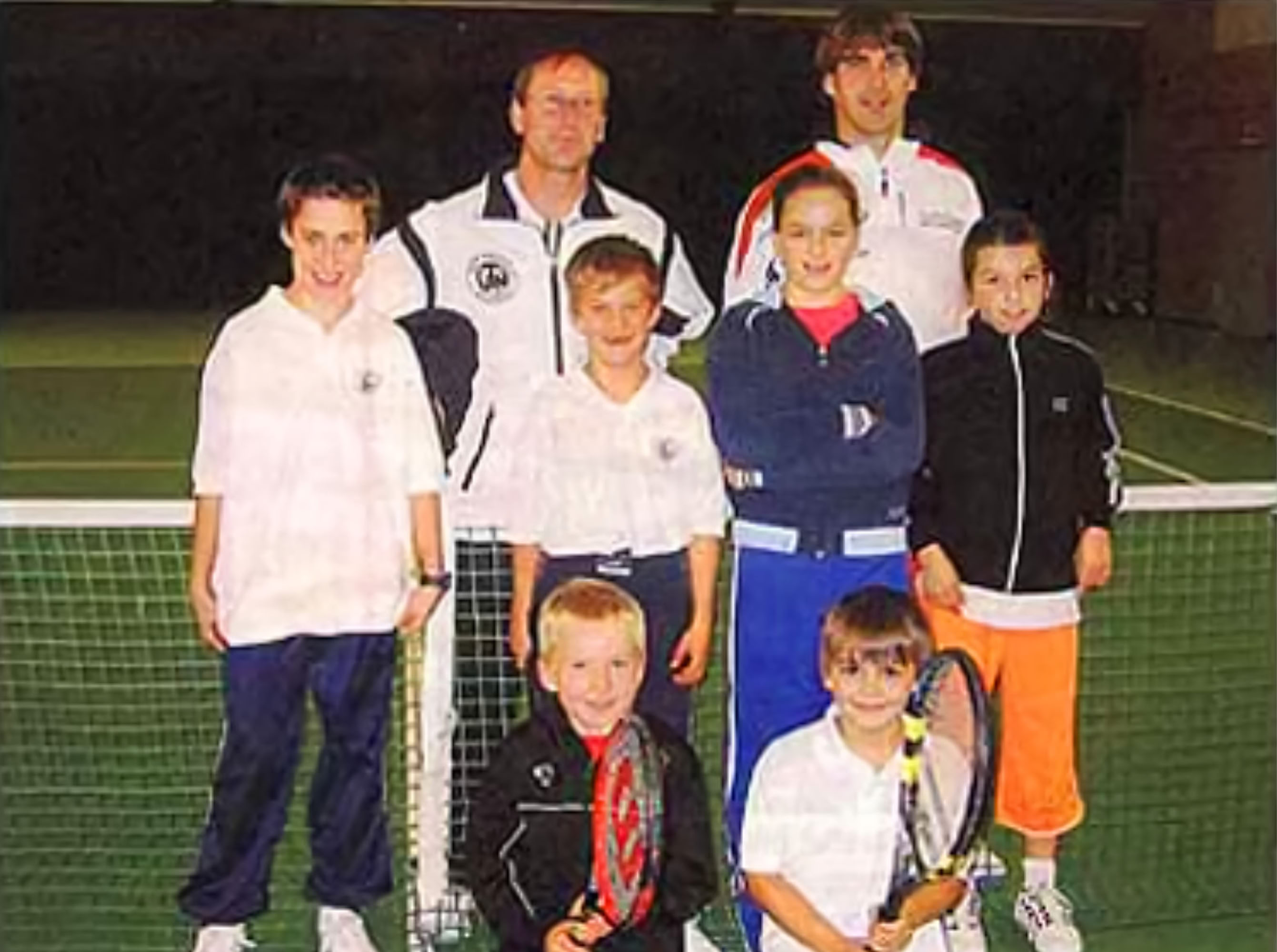 Eine Gruppe von sieben Personen, darunter zwei Erwachsene und f&uuml;nf Kinder, steht auf einem Tennisplatz vor einem Netz. Die Kinder halten Tennisschl&auml;ger.