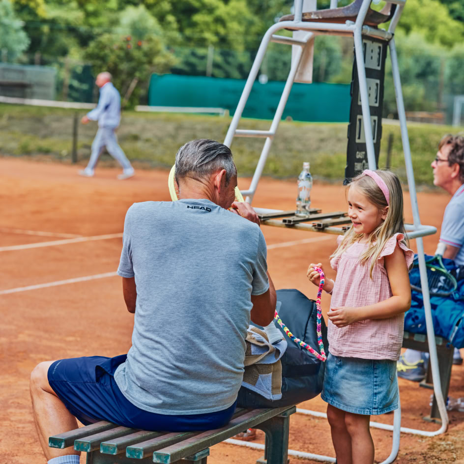 Ein Mann sitzt auf einer Bank neben einem Tennisplatz, spricht mit einem M&auml;dchen, das steht. Ein Schiedsrichterstuhl ist im Hintergrund zu sehen.