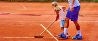 Ein Kind und ein Erwachsener stehen auf einem Tennisplatz aus Sand. Das Kind h&auml;lt einen Besen und zieht eine Linie im Sand. Der Erwachsene tr&auml;gt ein blaues Outfit.