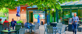 Ein Au&szlig;enbereich eines Caf&eacute;s mit mehreren Personen, die an Tischen sitzen oder stehen; im Hintergrund sind B&auml;ume und ein Geb&auml;ude mit Glasfront zu sehen.