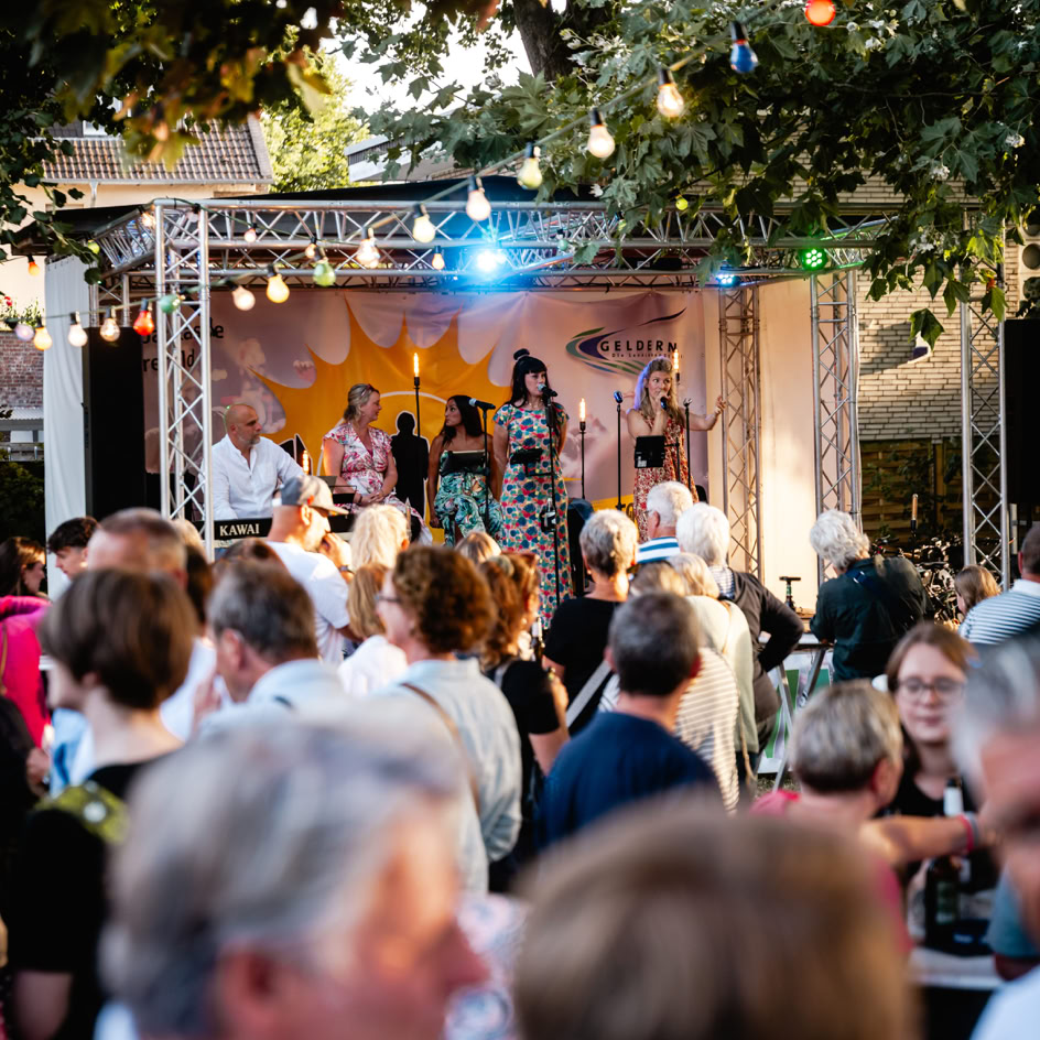 Eine Menschenmenge steht vor einer B&uuml;hne im Freien, auf der Musiker unter bunten Lichtern auftreten.