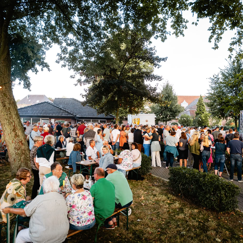 Eine gro&szlig;e Menschenmenge versammelt sich im Freien unter B&auml;umen, einige sitzen an Tischen, w&auml;hrend andere stehen. Im Hintergrund sind Geb&auml;ude und ein klarer Himmel zu sehen.