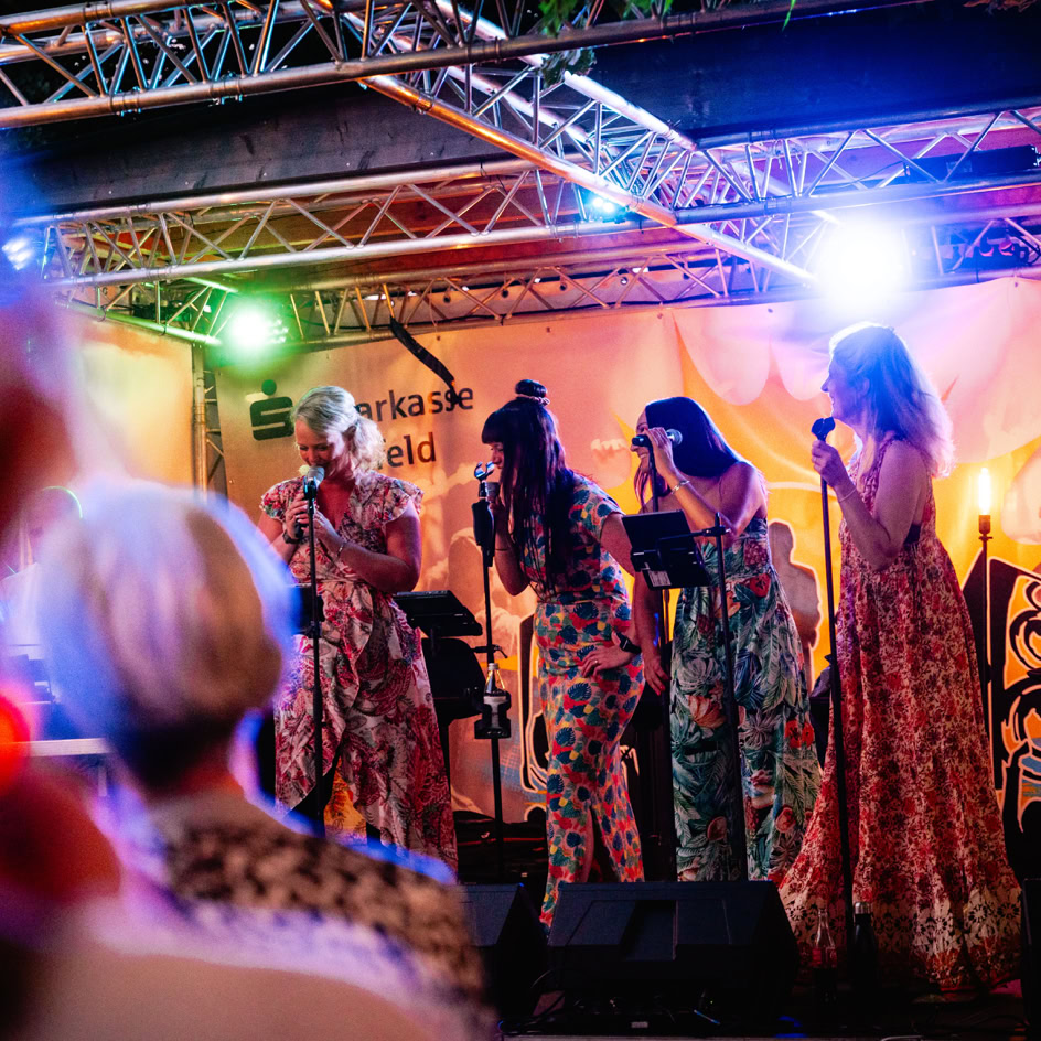 Vier Personen singen auf einer B&uuml;hne mit bunten Lichtern und Mikrofonen, Publikum im Vordergrund.