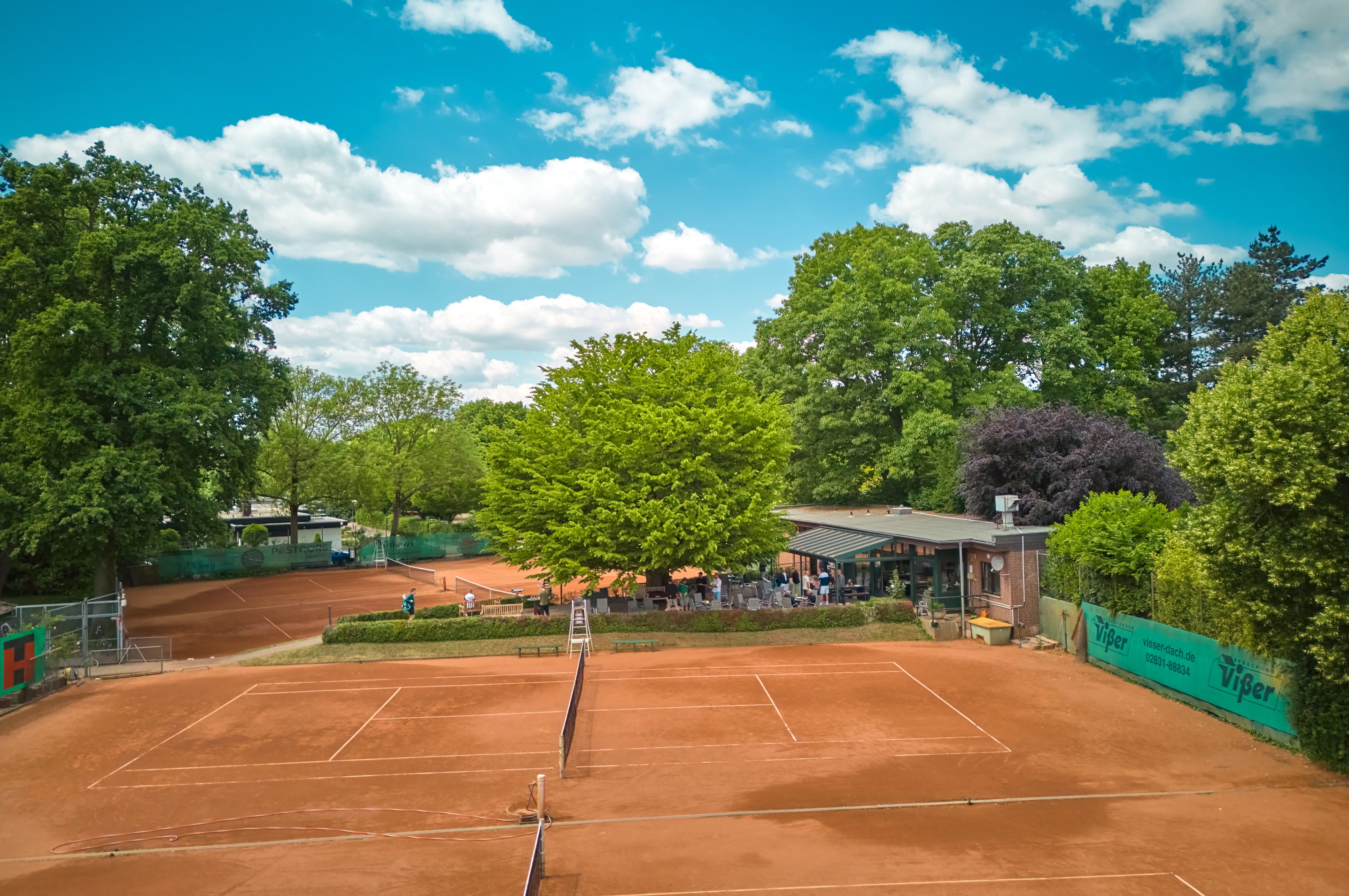 Zwei Sandtennispl&auml;tze umgeben von B&auml;umen, mit einem Geb&auml;ude im Hintergrund unter blauem Himmel mit Wolken.