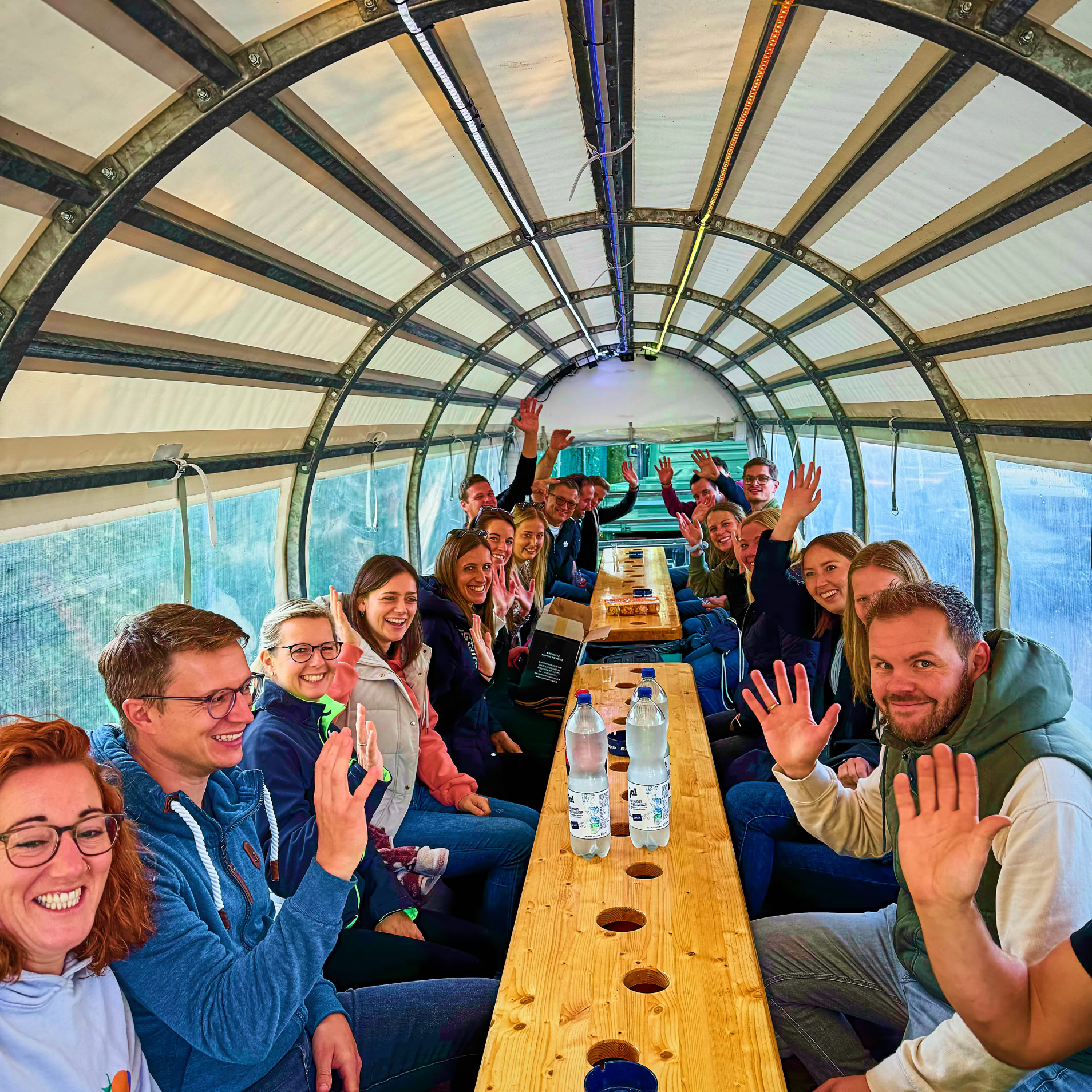 Personengruppe sitzt an einem langen Tisch in einem &uuml;berdachten Bereich und winkt in die Kamera.