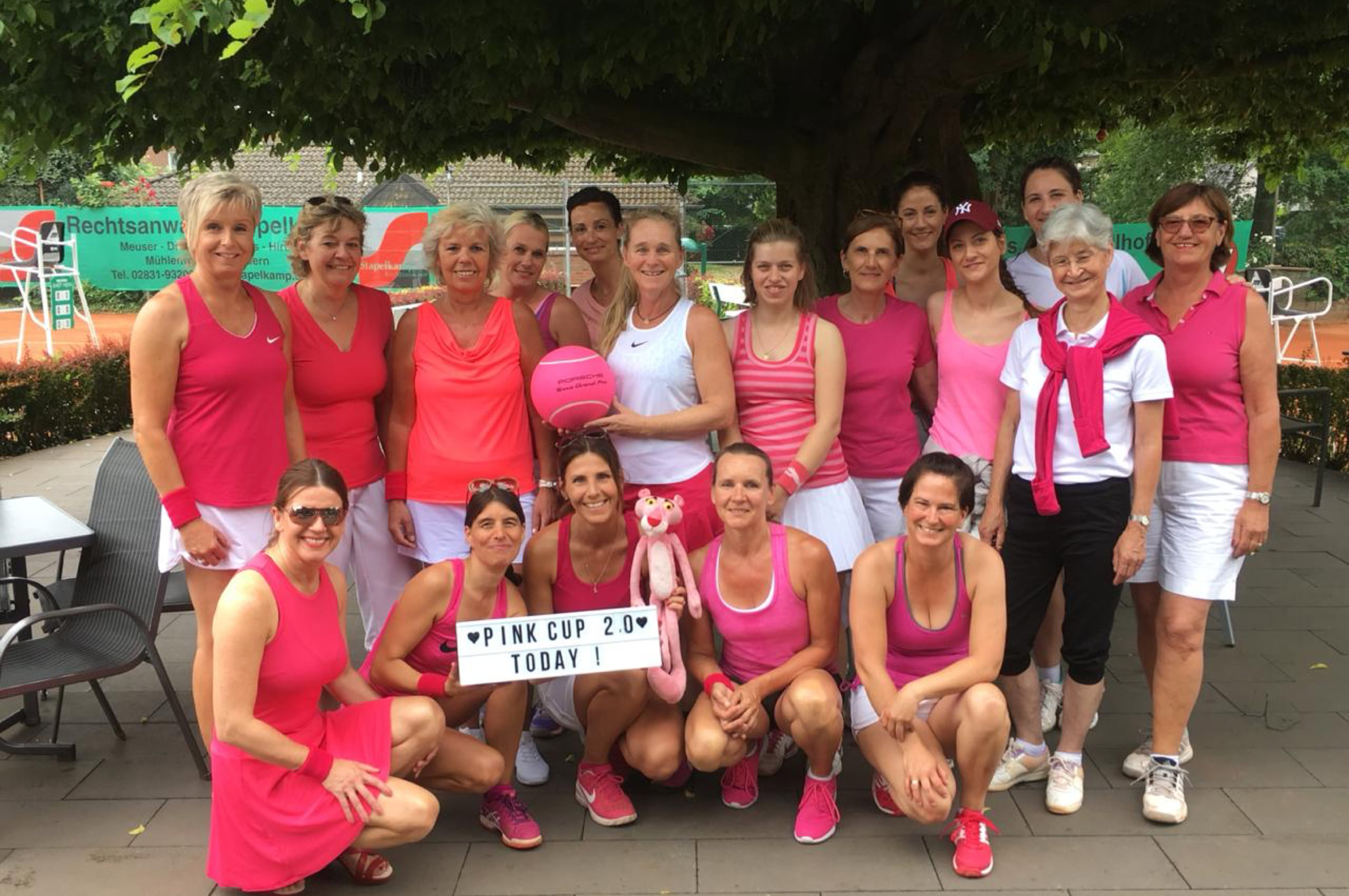 Gruppe von Frauen in pinker Sportkleidung posiert drau&szlig;en, einige halten ein Schild und einen Ball.