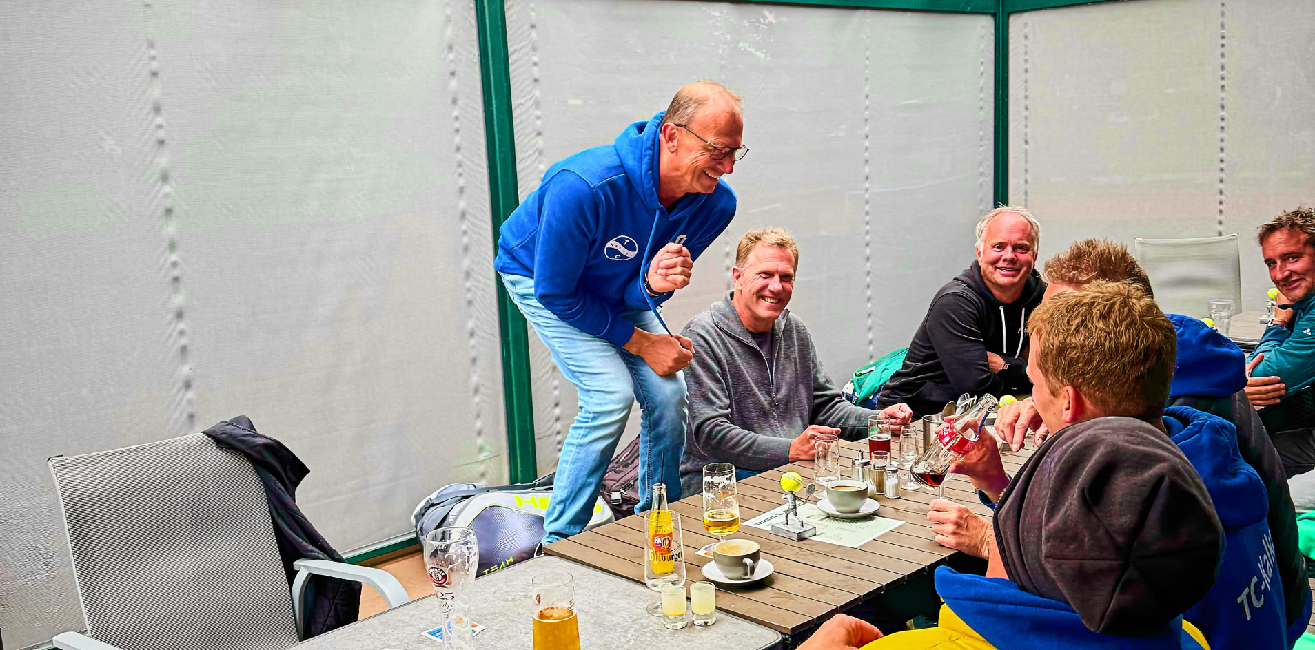 Eine Gruppe von Menschen sitzt an einem Tisch unter einem Glasdach, einige lachen und trinken.