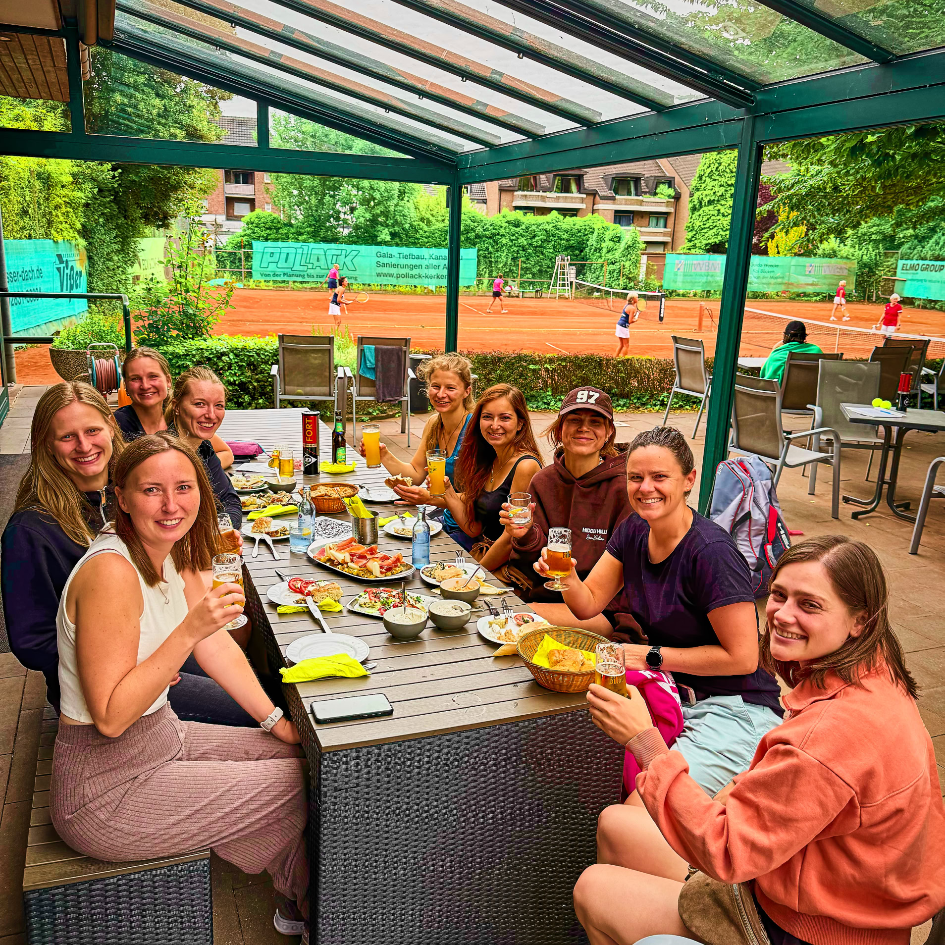 Eine Gruppe von Menschen sitzt an einem Tisch im Freien unter einem Glasdach, mit Essen und Getr&auml;nken auf dem Tisch. Im Hintergrund sind B&auml;ume und ein Tennisplatz zu sehen.