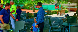 Eine Gruppe von Menschen steht und unterh&auml;lt sich auf einer Terrasse mit Tischen und St&uuml;hlen, im Hintergrund sind B&auml;ume und ein Tennisplatz zu sehen.