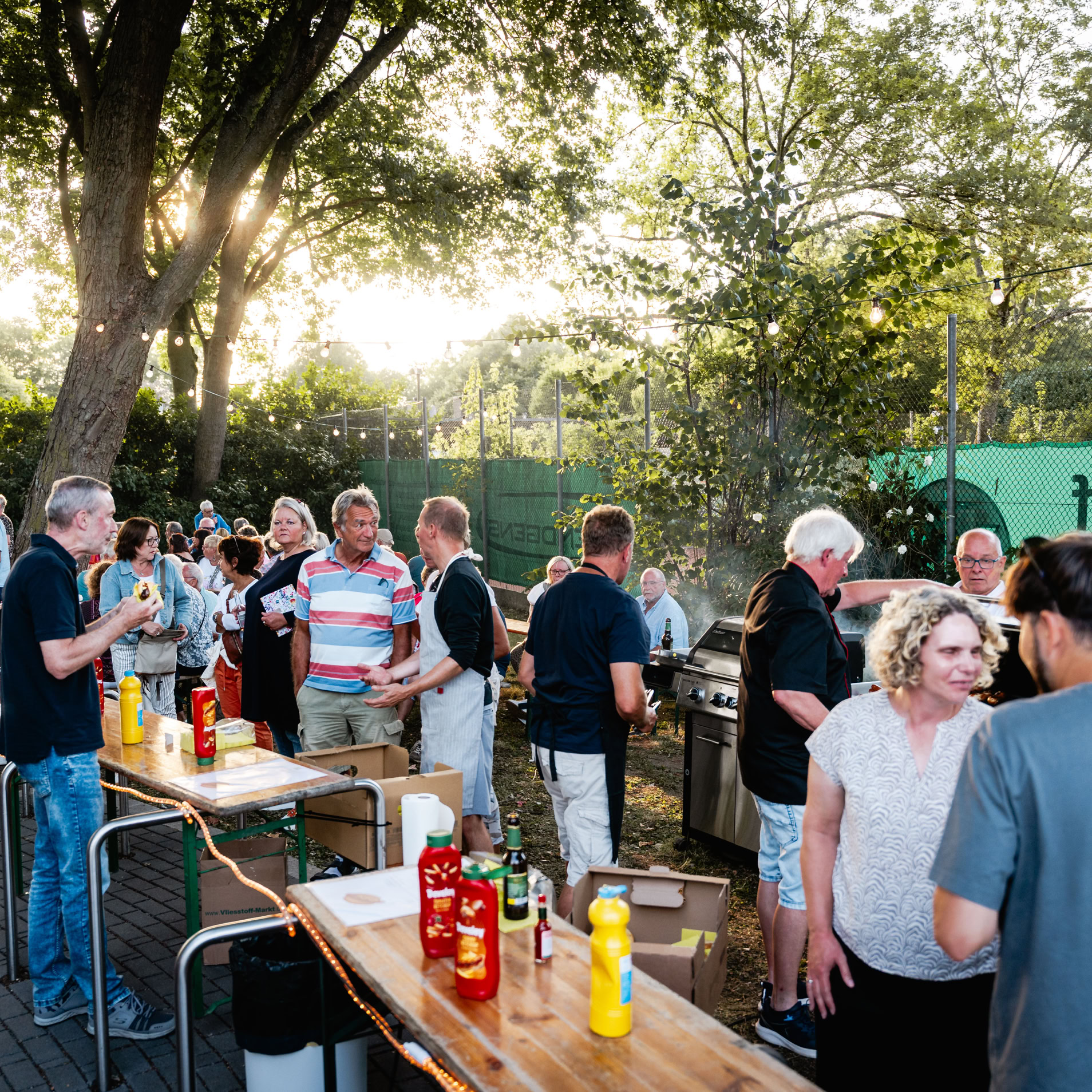 Menschen stehen bei einem Outdoor-Event um Tische mit Ketchup- und Senfflaschen, im Hintergrund B&auml;ume und ein gr&uuml;ner Zaun.