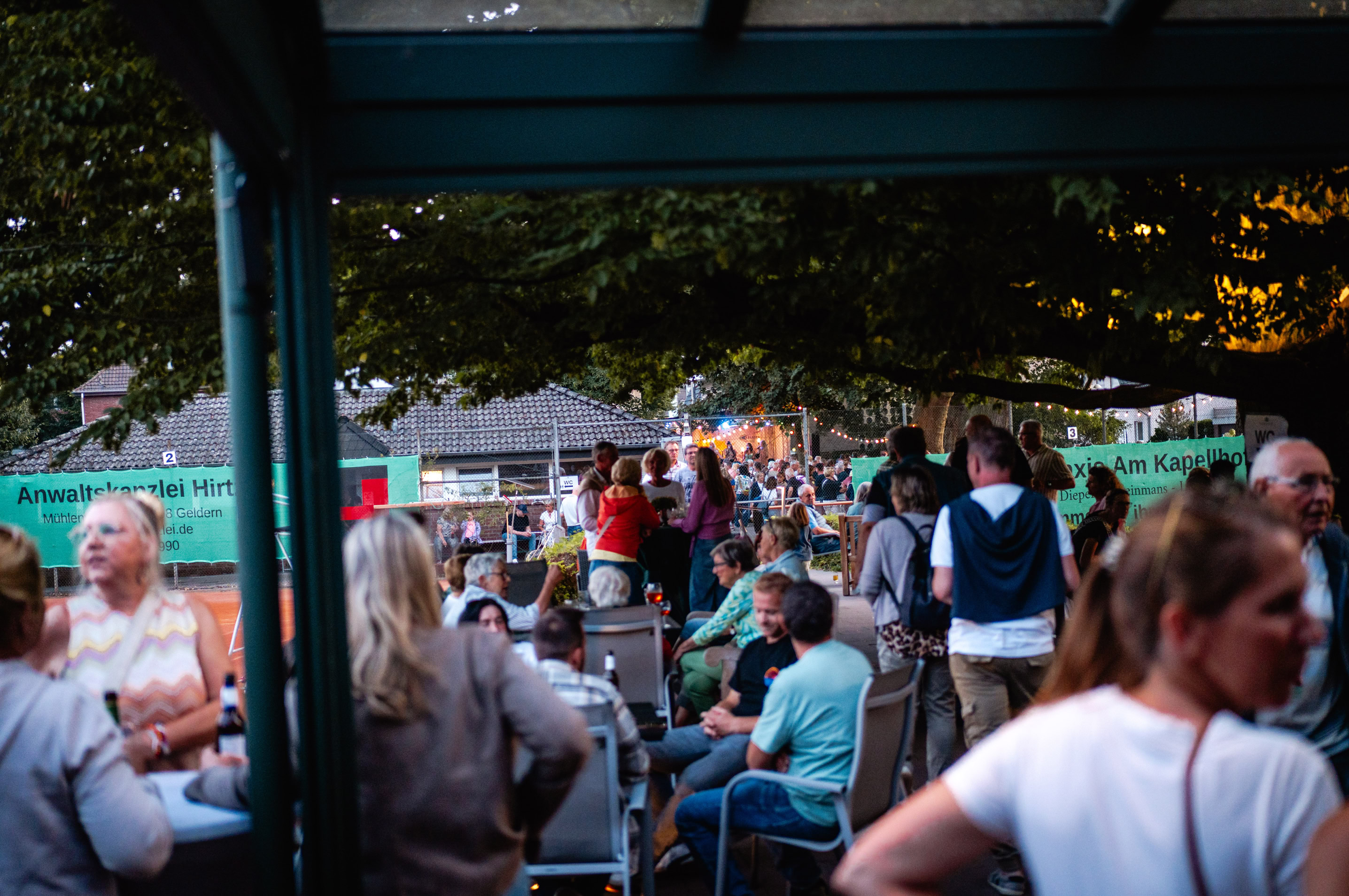 Eine Gruppe von Menschen versammelt sich im Freien unter einem &uuml;berdachten Bereich, einige sitzen, andere stehen, umgeben von B&auml;umen und einem Geb&auml;ude im Hintergrund.