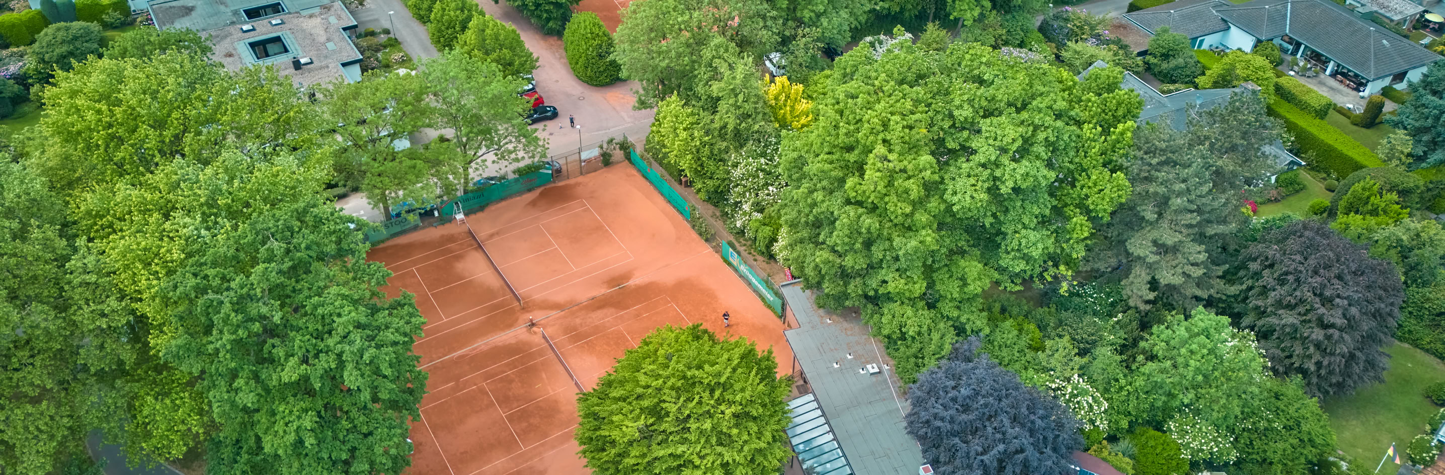 Luftaufnahme von mehreren Tennispl&auml;tzen mit rotem Sand, umgeben von B&auml;umen und Geb&auml;uden.