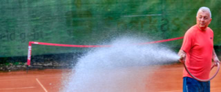 Eine Person in einem roten T-Shirt bew&auml;ssert einen Tennisplatz mit einem Schlauch.